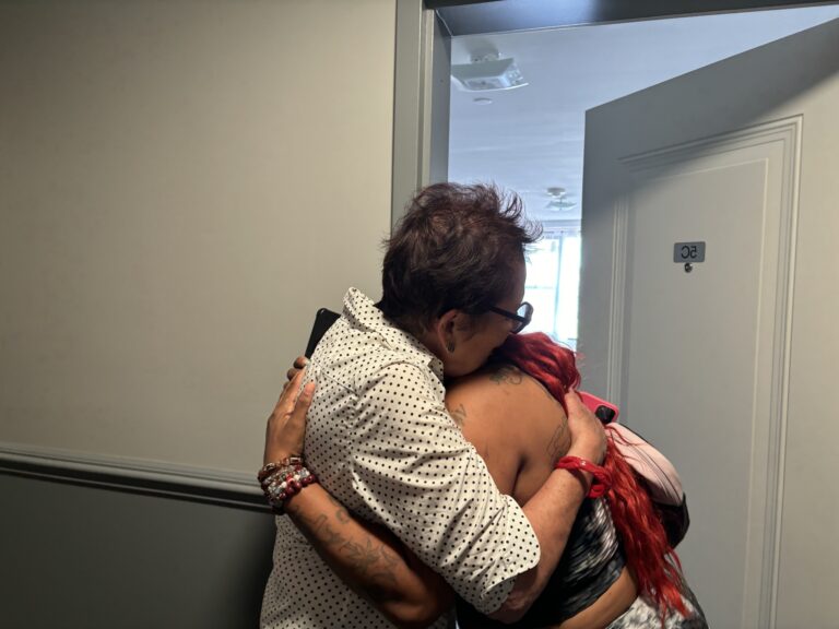 Two people embrace in a doorway. One person has red hair the other wears a polka dot shirt.