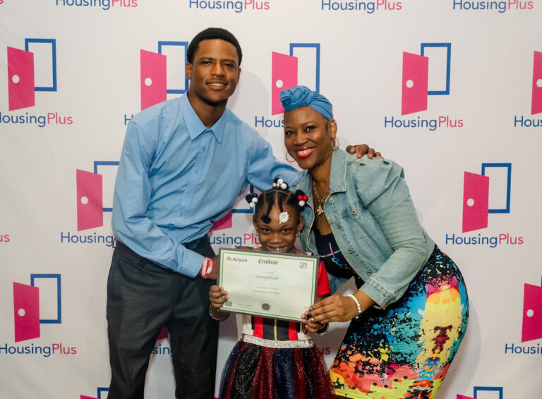 Three people stand in front of a sign reading "HousingPlus" there is a young girl in the center and two adults behind her.