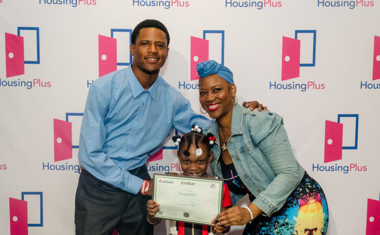 Three people stand in front of a sign reading "HousingPlus" there is a young girl in the center and two adults behind her.