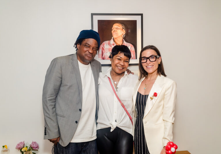 Three people stand in front of a framed portrait of a woman closing her eyes.