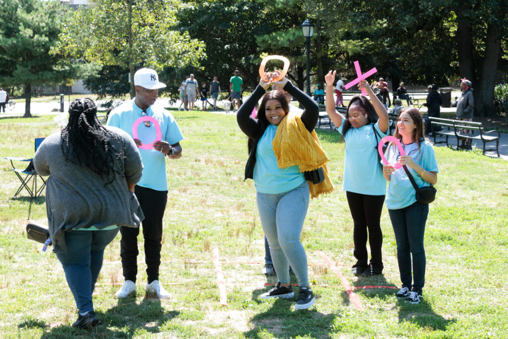 Four people stand in bright blue shirts in a green field of grass. They are playing life sized tic tac toe.