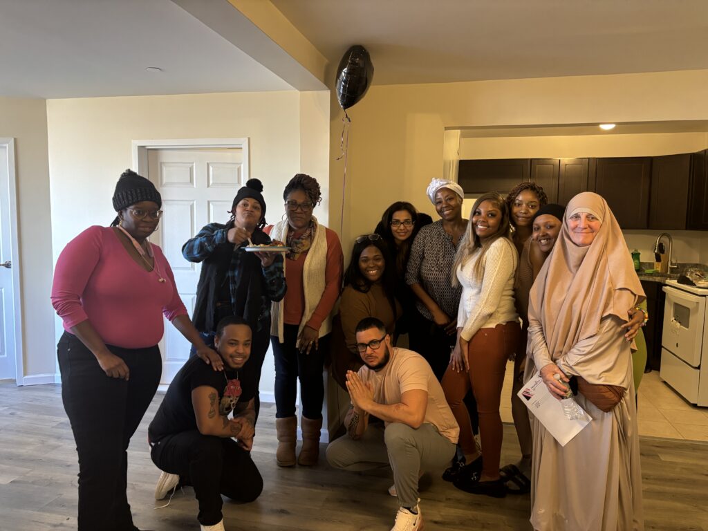 A group of individuals pose and smile at camera. There is a woman in a head covering on the right side, and then there are several women clustered together. A kitchen is behind them.