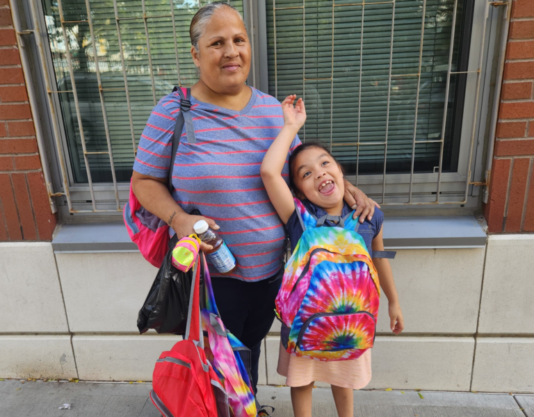 A smiling little girl raises her hand in the air. She is wearing a tye-die backpack. Beside her, her grandmother holds another red backpack and smiles. They are outdoors in front of a building.