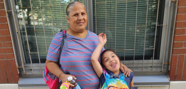 A smiling little girl raises her hand in the air. She is wearing a tye-die backpack. Beside her, her grandmother holds another red backpack and smiles. They are outdoors in front of a building.