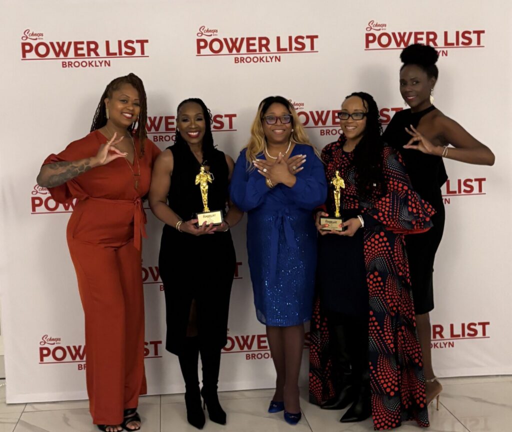 Four African American women stand in front of a step and repeat with the word Power List visible. They make the symbol for Zeta sorority.
