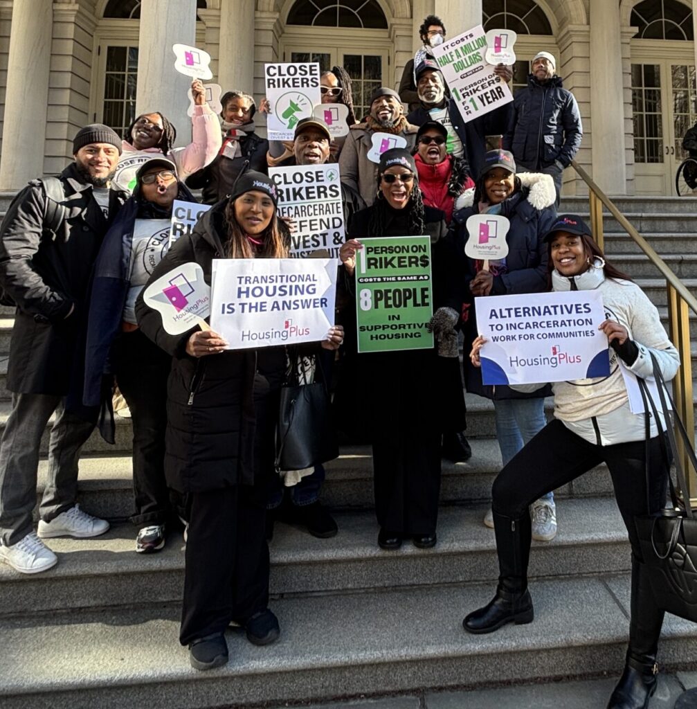 A group of HousingPlus staff stand on the steps of City Hall with signs advocating for the closure of Rikers Island.