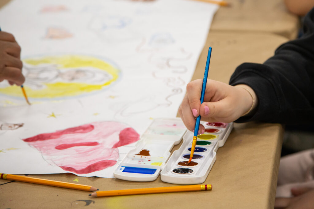 Shows two hands painting on a white sheet of paper, with a yellow circular form and several paints visible in the frame.